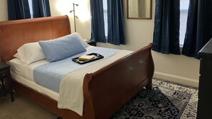 A neatly made bed with a wooden headboard, a bedside table, and a nightstand. There is a patterned rug on the floor, and the room has a large mirror on the wall.