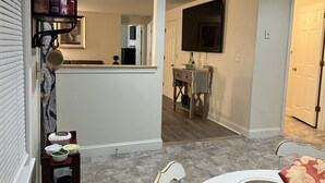 A modern kitchen with a central island, a dining area with a white table and chairs, and a hallway leading to a bedroom.