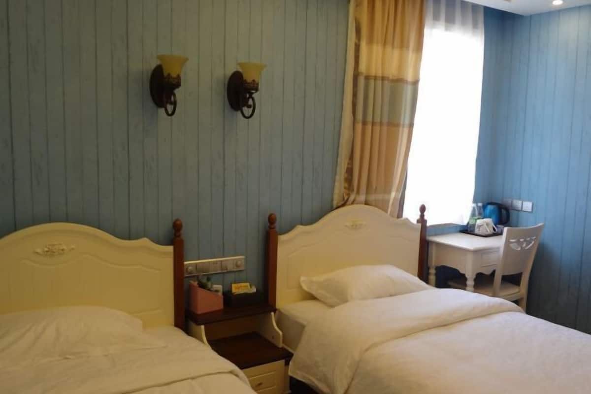 A hotel room with two single beds, a small desk with a chair, a nightstand with a lamp, and a window with curtains.