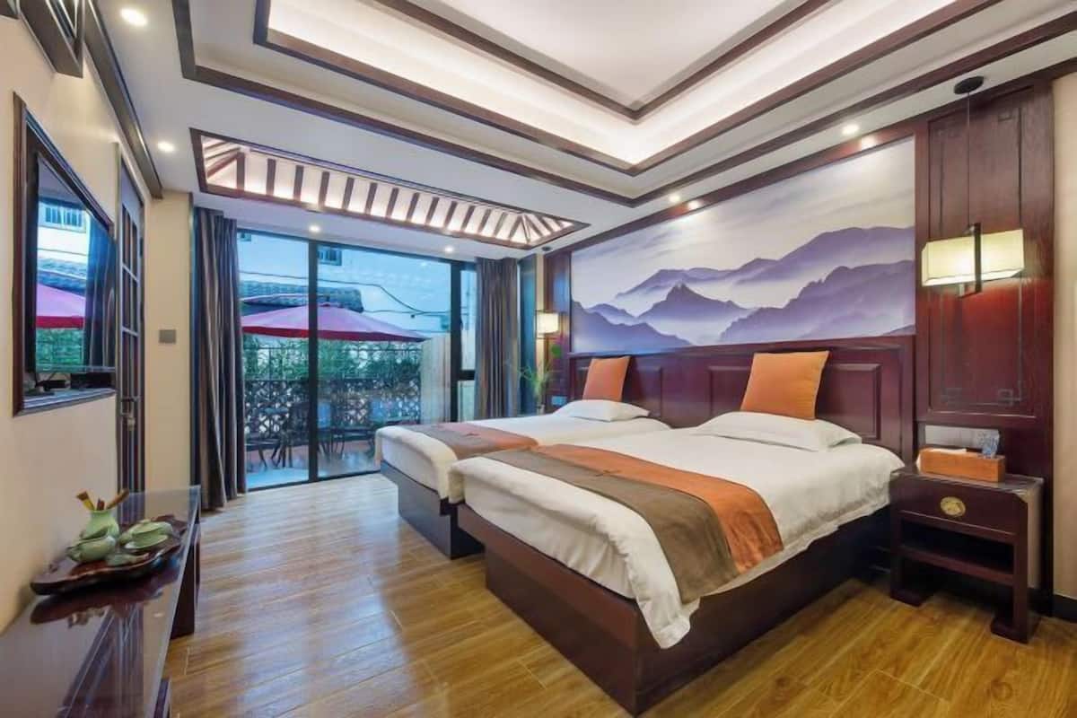A hotel room with a large bed, wooden flooring, a painting of mountains, and a view of the outdoors.