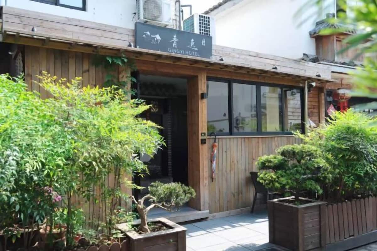A wooden exterior with a sign for 'Ginghi Hotel.' There are potted plants and a wooden fence in the foreground.