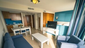 Un salon moderne avec un canapé, des fauteuils, une table basse, un téléviseur et une cuisine en arrière-plan.