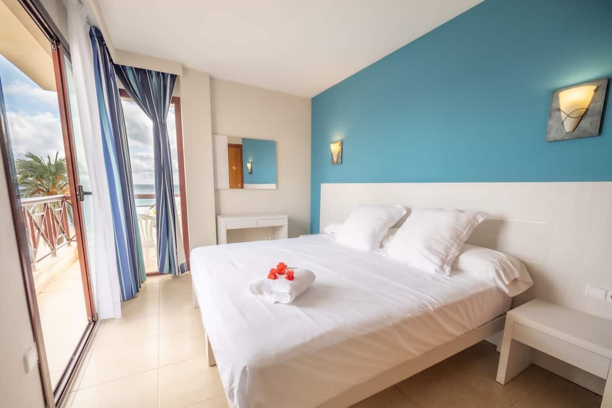 A hotel room with a bed, a nightstand, a desk, and a view of the ocean through a sliding door.