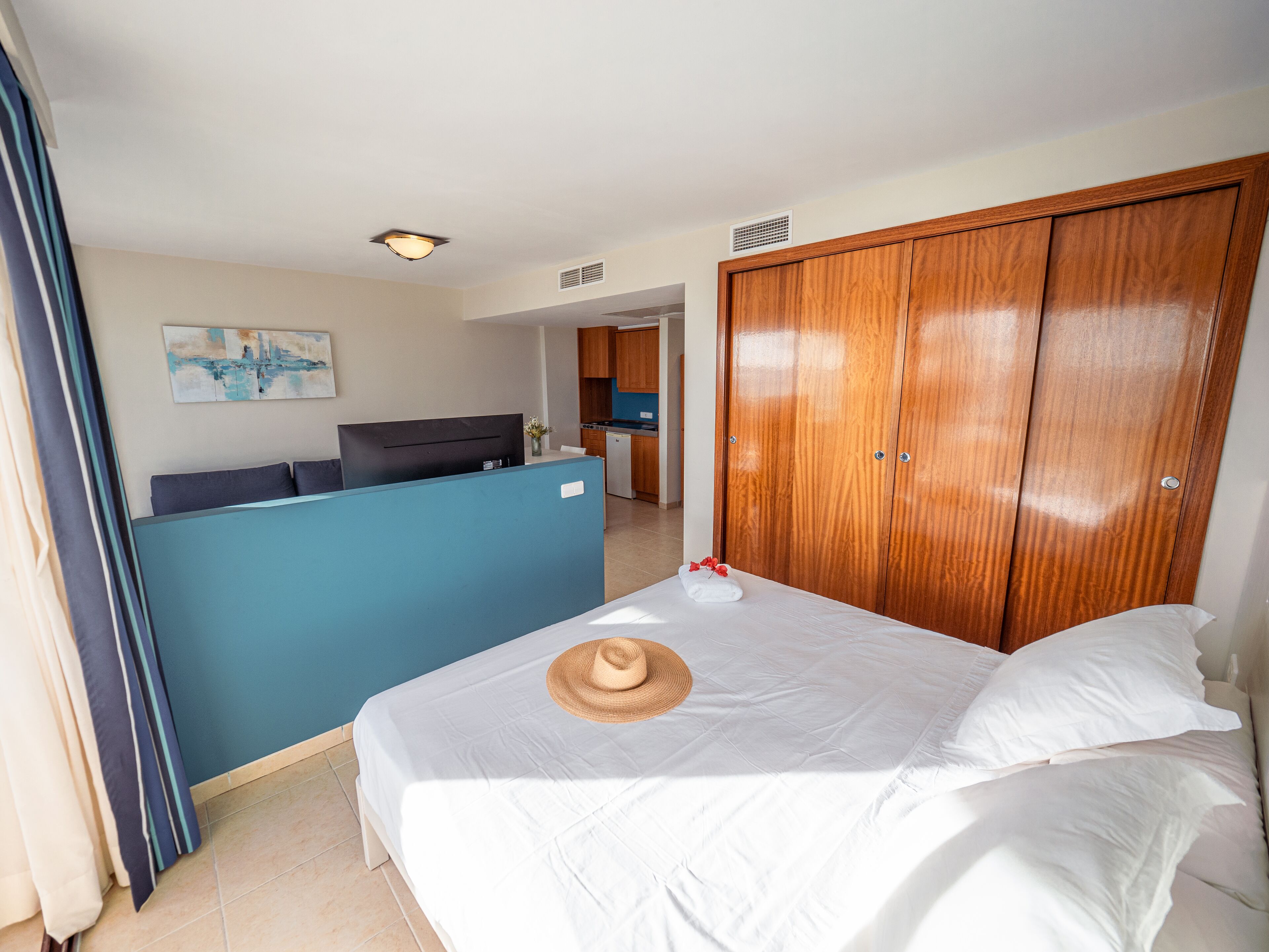 A hotel room with a bed, a wooden wardrobe, a desk with a computer, and a view of the kitchen.