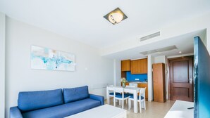 Un salon moderne avec un canapé bleu, une table basse blanche et un coin cuisine doté de meubles en bois et d’un plan de travail bleu.