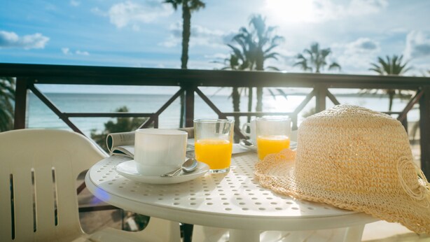 Une table avec un chapeau blanc, deux tasses et deux verres de jus d’orange, sur un balcon offrant une vue sur l’océan et les palmiers.