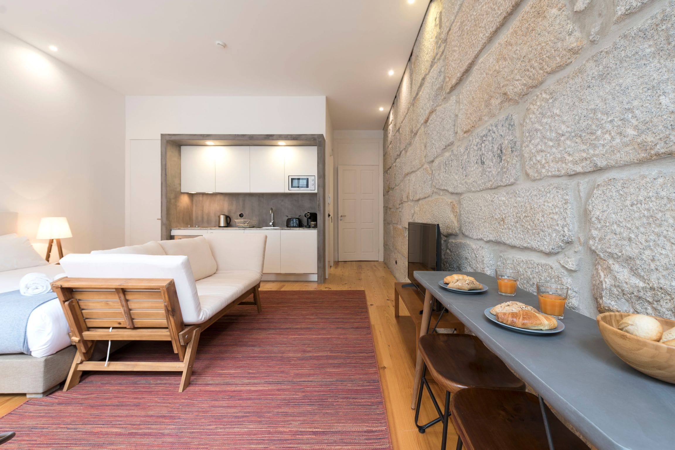A compact hotel room with a kitchenette, a bed, a dining area with breakfast, and a stone wall.