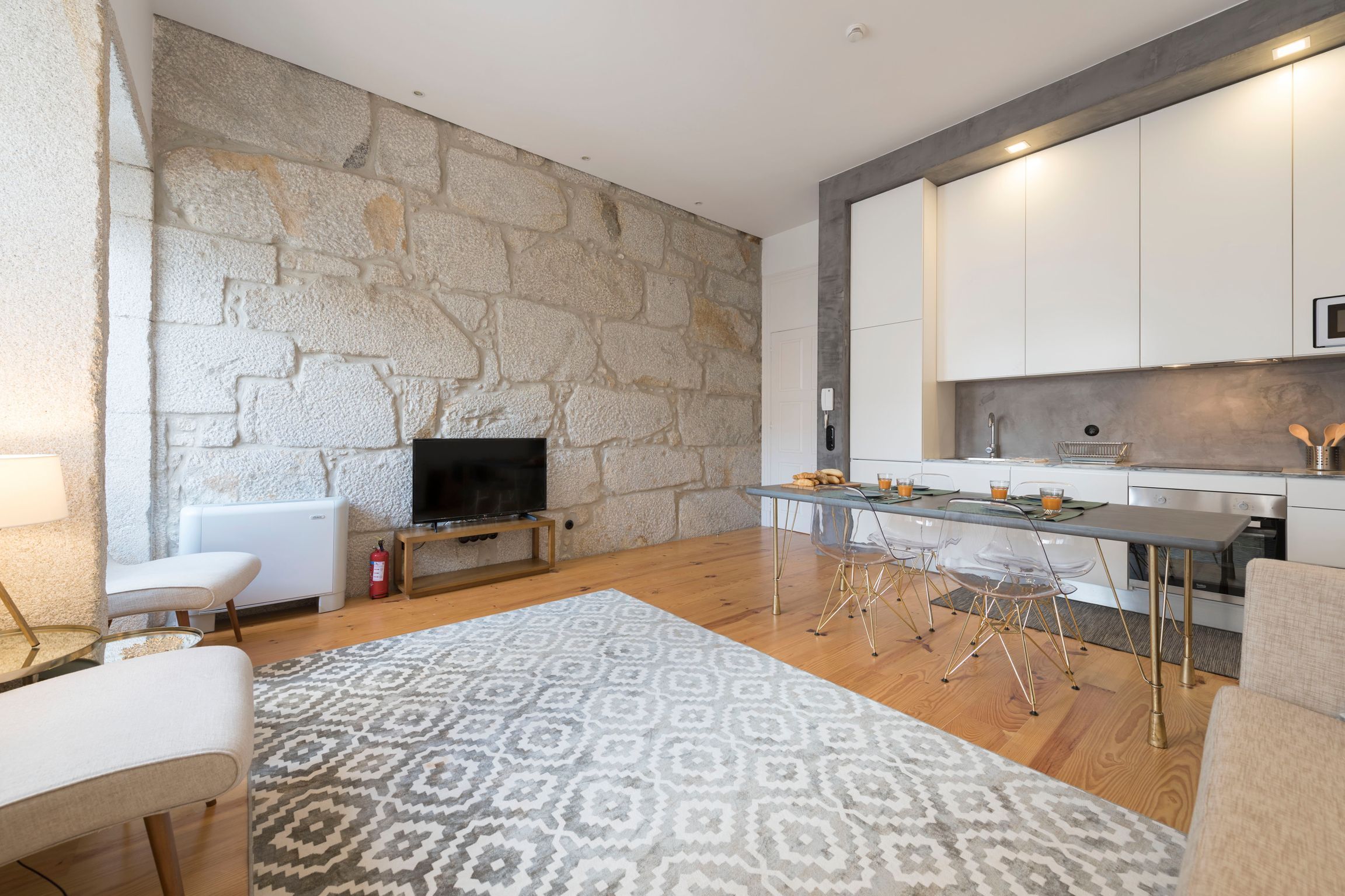 A modern living room with a stone wall, a flat-screen TV, a dining area with a glass table, and a kitchen with white cabinets.