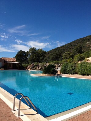 Une piscine d’un bleu éclatant, bordée de pierres, avec un escalier en métal, et offrant une vue sur une colline parsemée d’arbres et sur un bâtiment en arrière-plan.