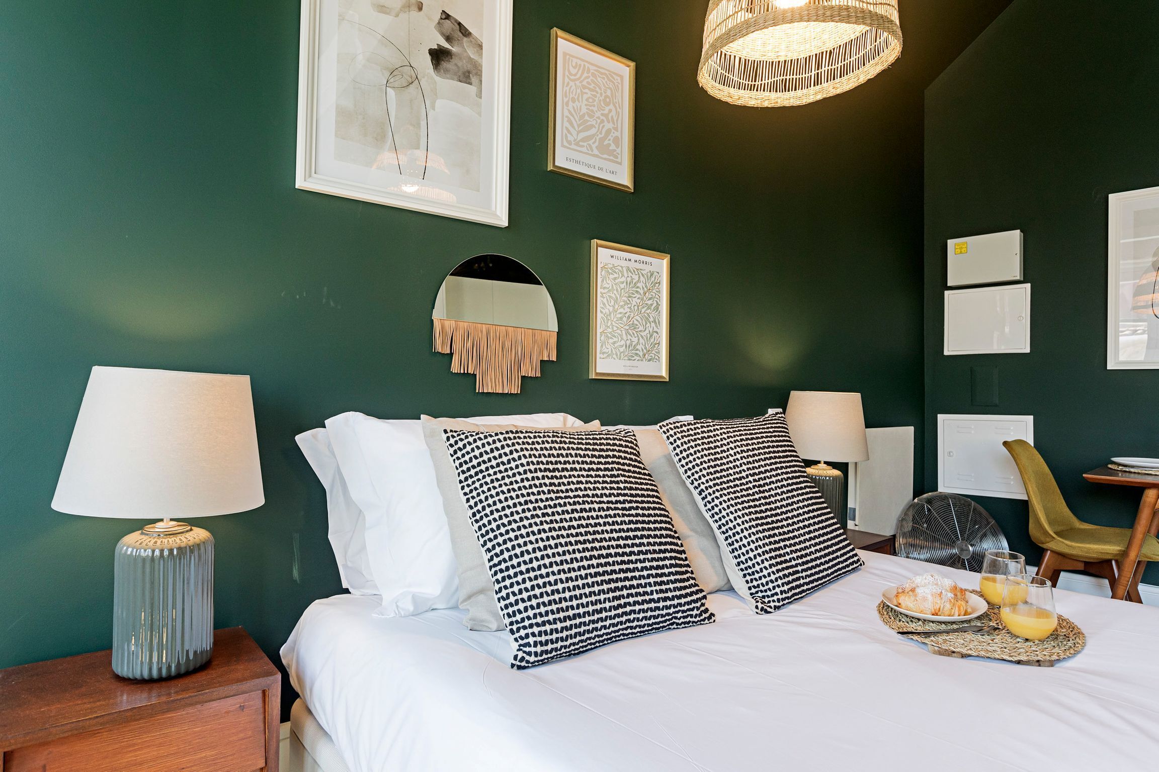 Hotelkamer met een bed, nachtlampen, een spiegel, kunstwerken in lijsten, een ventilator, een stoel en een ontbijtplateau met sinaasappelsap.