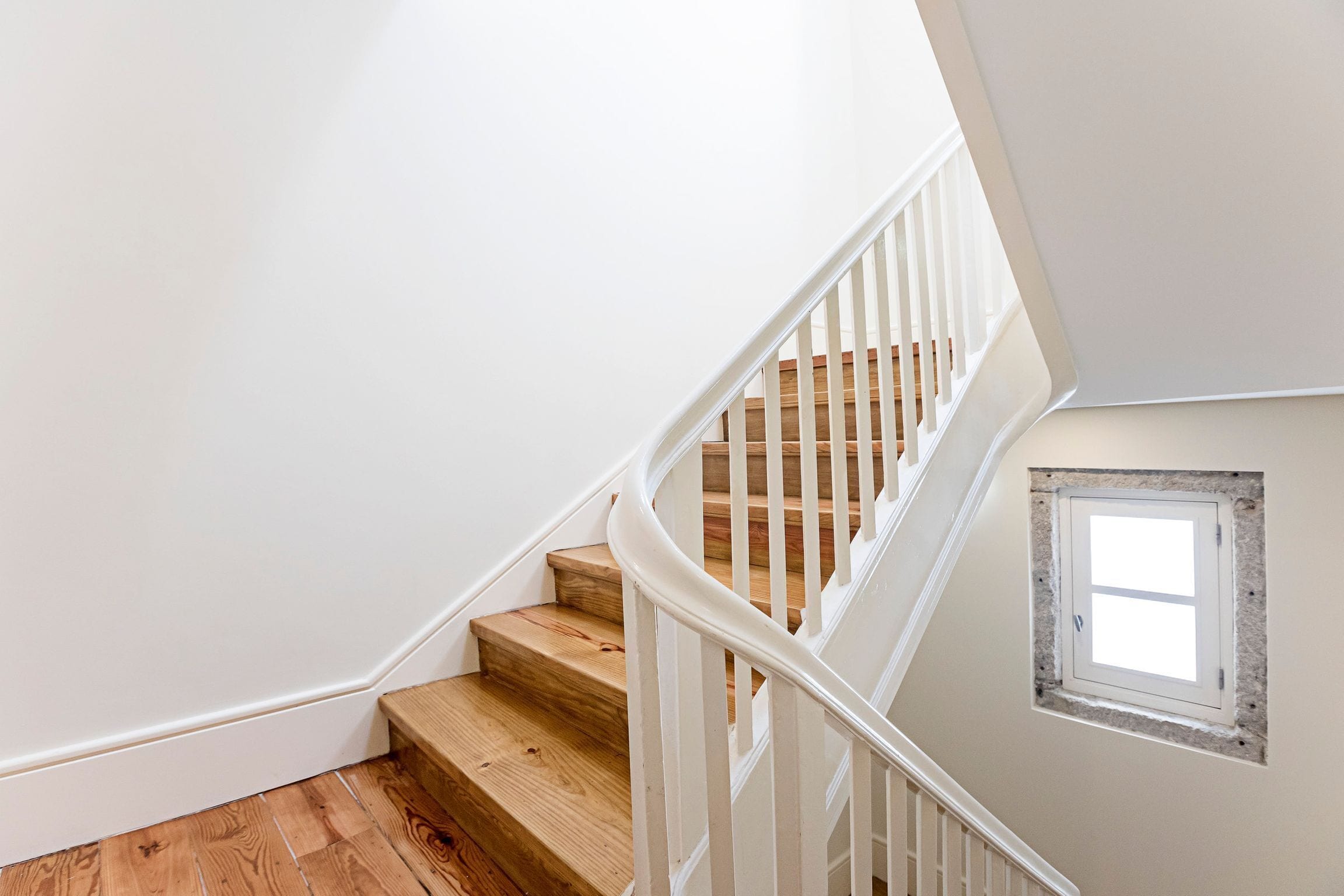 Un escalier avec des marches en bois et une rampe blanche, menant à un palier doté d’une petite fenêtre.