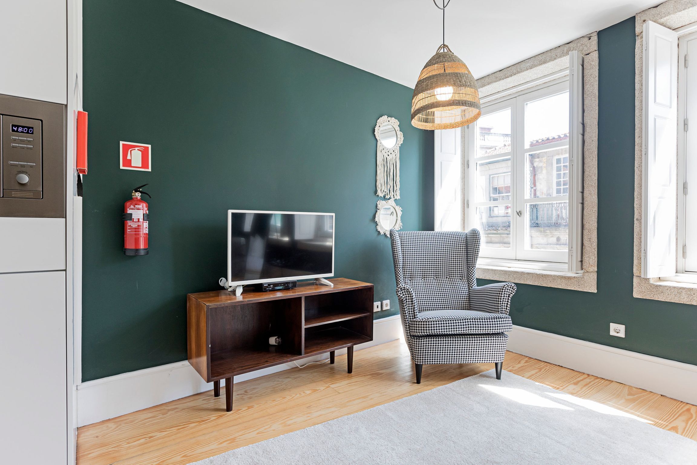 A living room with a television on a wooden stand, a checkered armchair, a hanging light fixture, and a fire extinguisher on the wall.