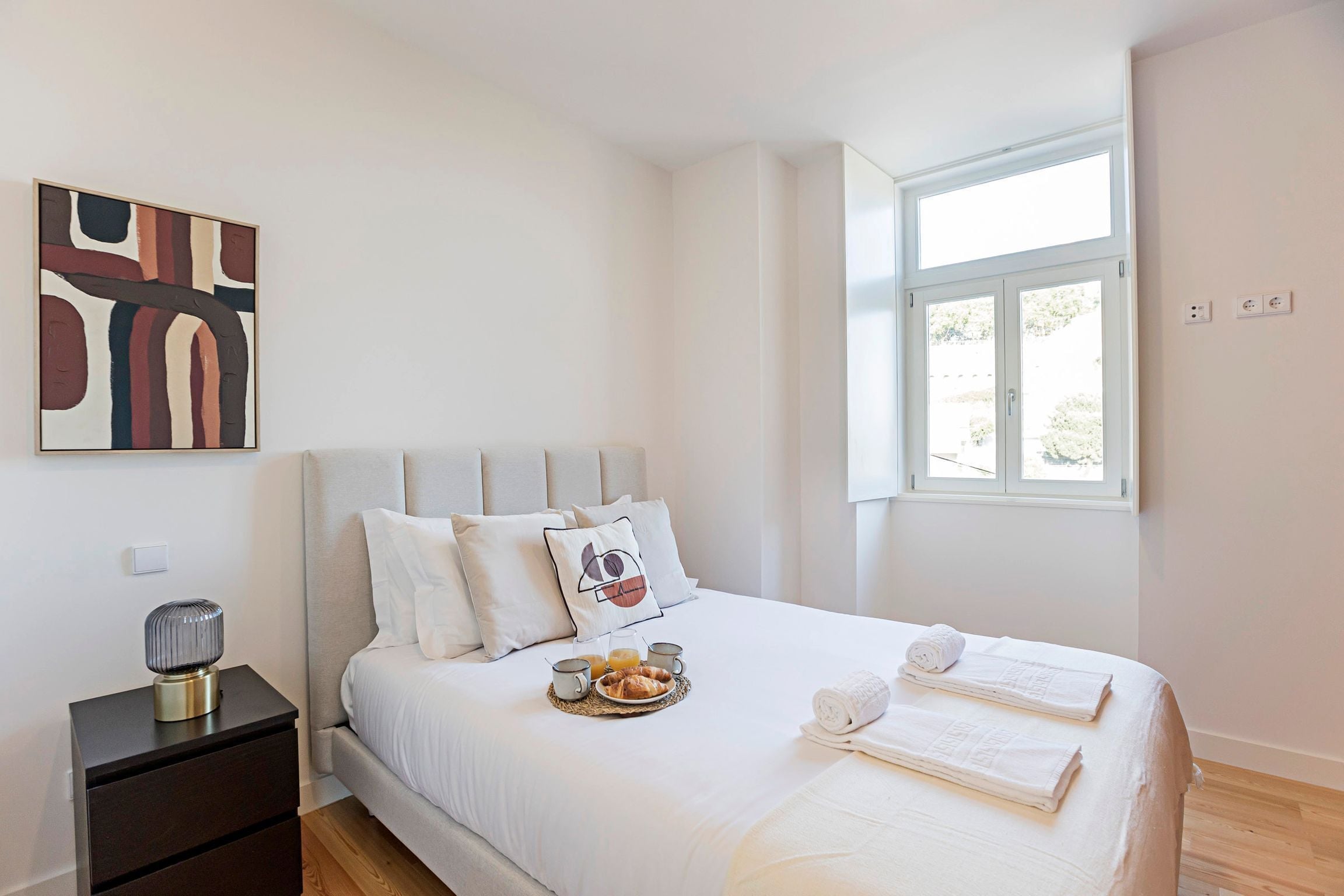 A modern hotel room with a bed, a bedside table with a lamp, a tray with breakfast, and a large window.
