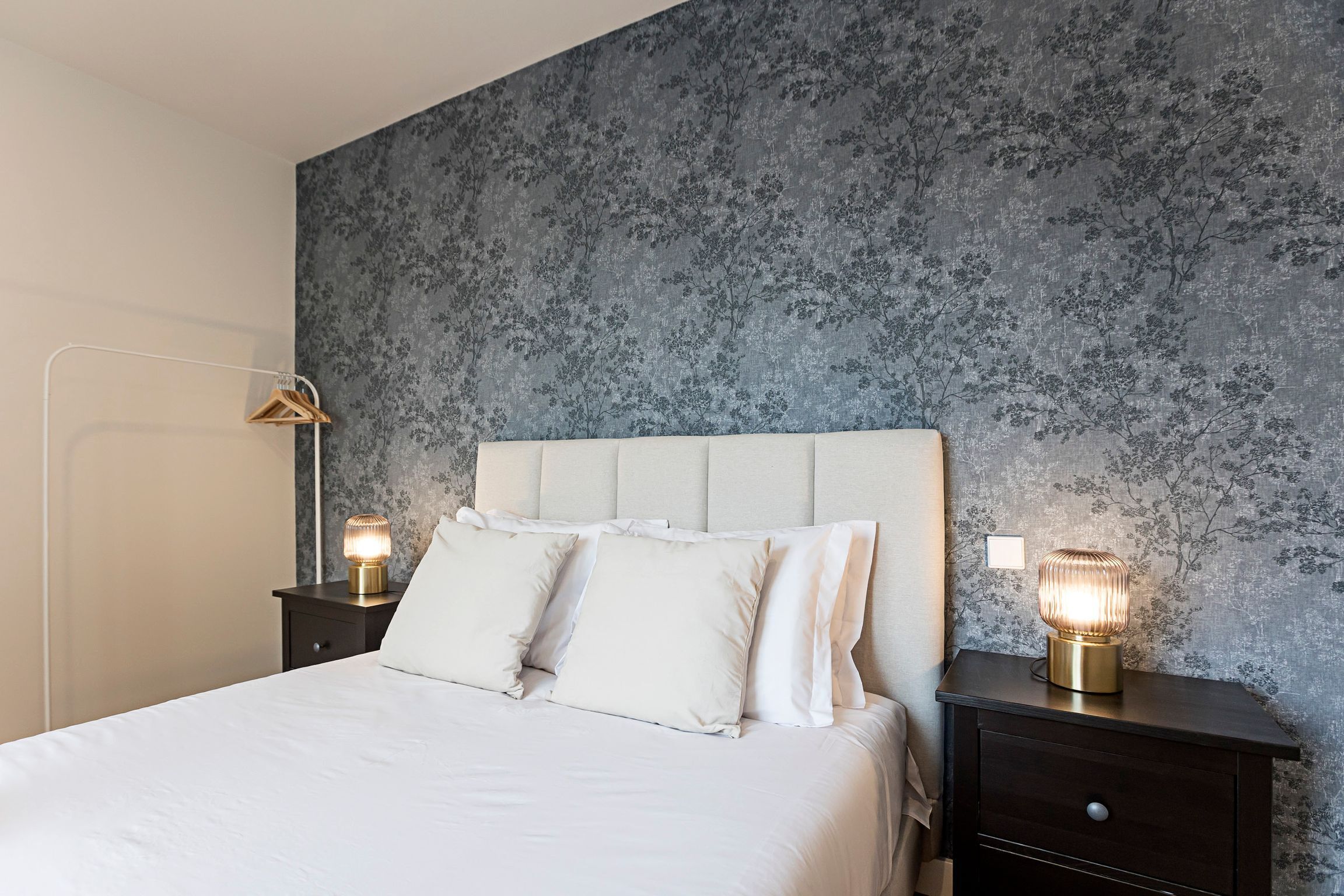 A neatly made bed with white linens and pillows, a dark wooden nightstand with a lamp, and a wall with a floral pattern.