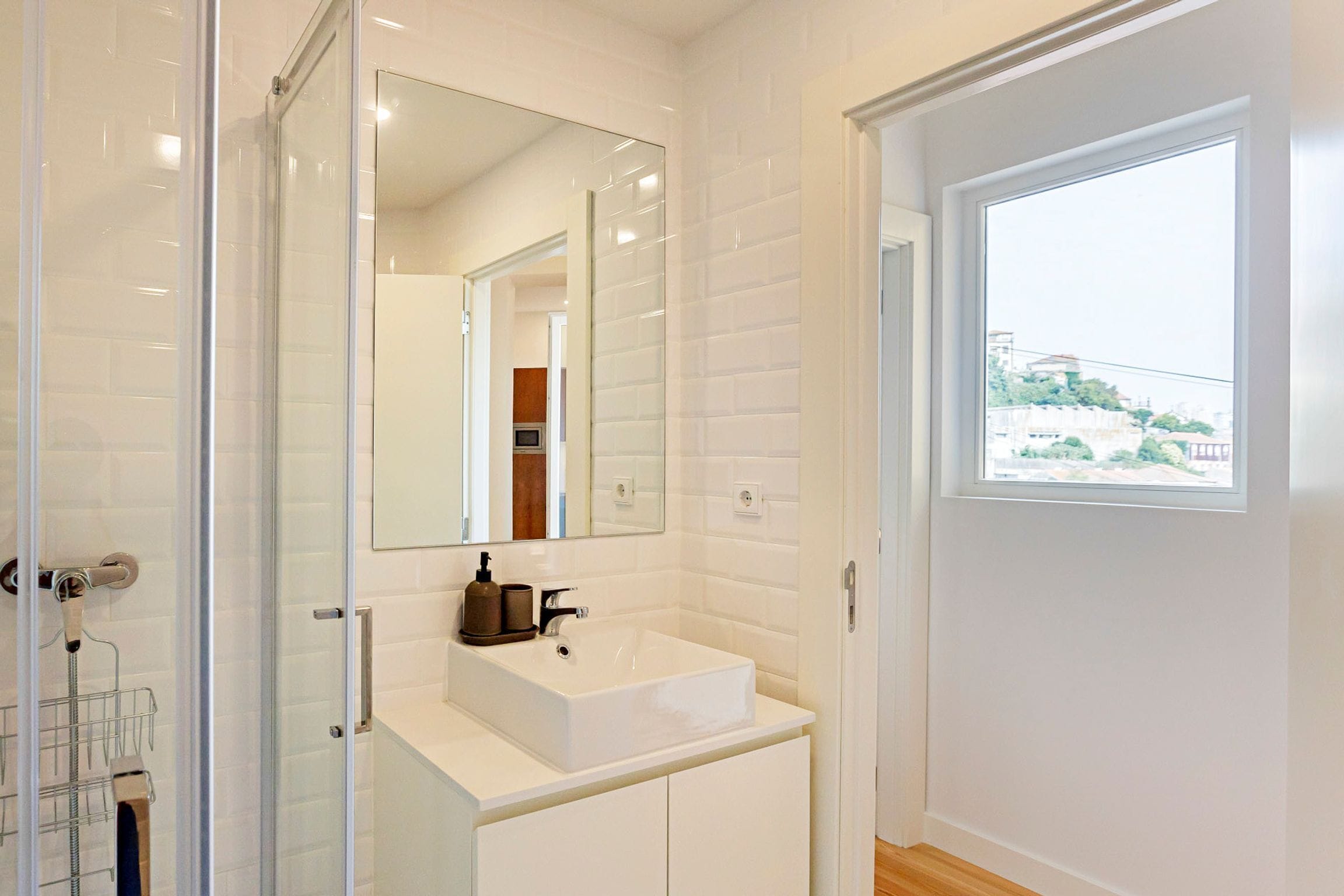 A modern bathroom with a glass shower enclosure, a white sink with a mirror above it, and a window providing a view of the outside.