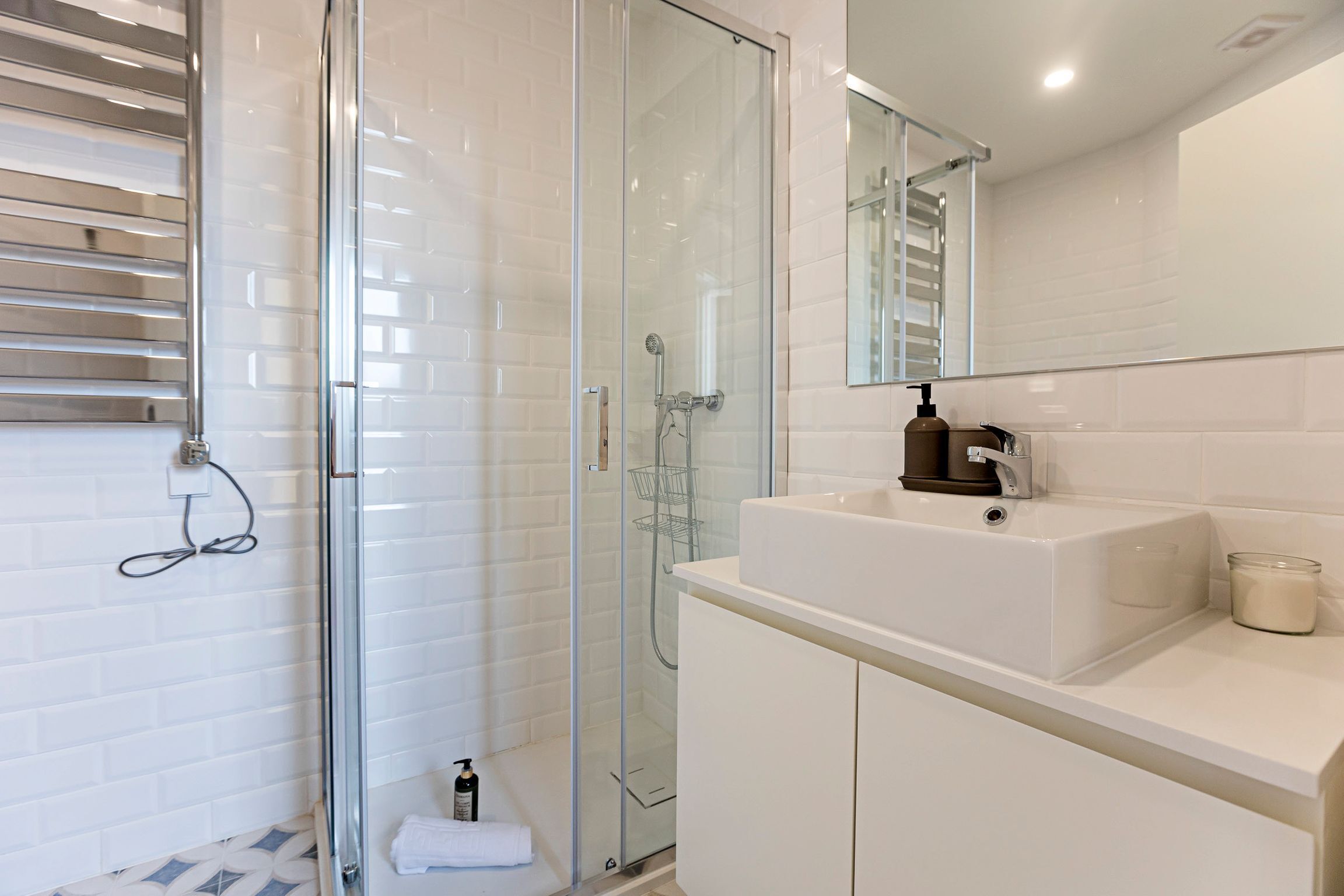 A modern bathroom with a glass-enclosed shower, white tiled walls, a white sink with a mirror above, and a small towel rack.