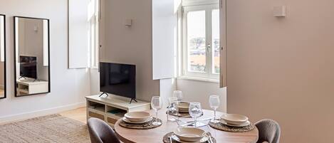 A dining area with a round table set for four, surrounded by chairs. A television is mounted on the wall to the left.