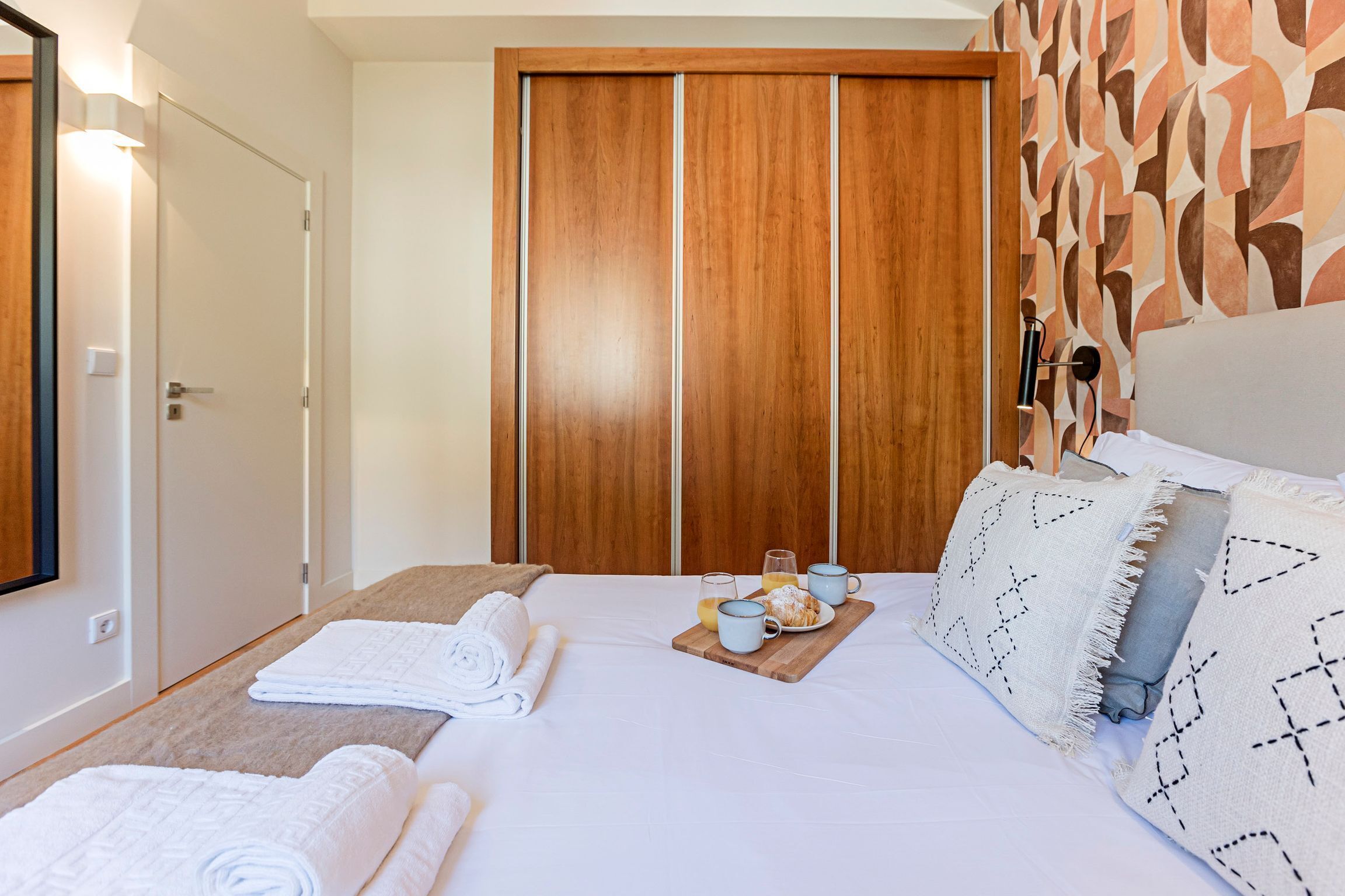A hotel room with a bed, two bedside tables, a wooden wardrobe, and a breakfast tray with cups and pastries.