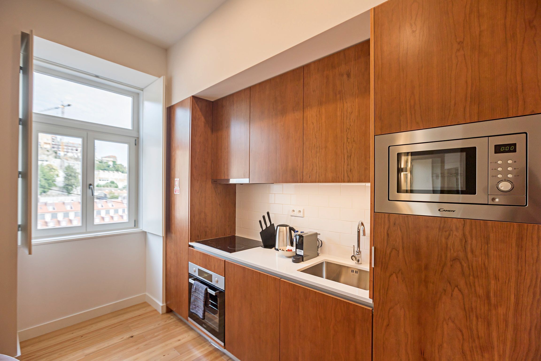 A modern kitchen with wooden cabinets, a built-in microwave, and a window with a view of buildings outside.