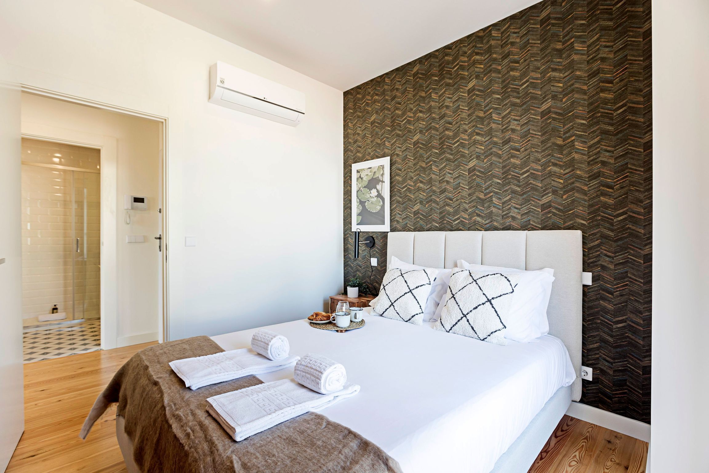 A neatly made bed with white linens and a brown throw blanket, a bedside table with a cup and a book, and a patterned wallpaper on the wall.