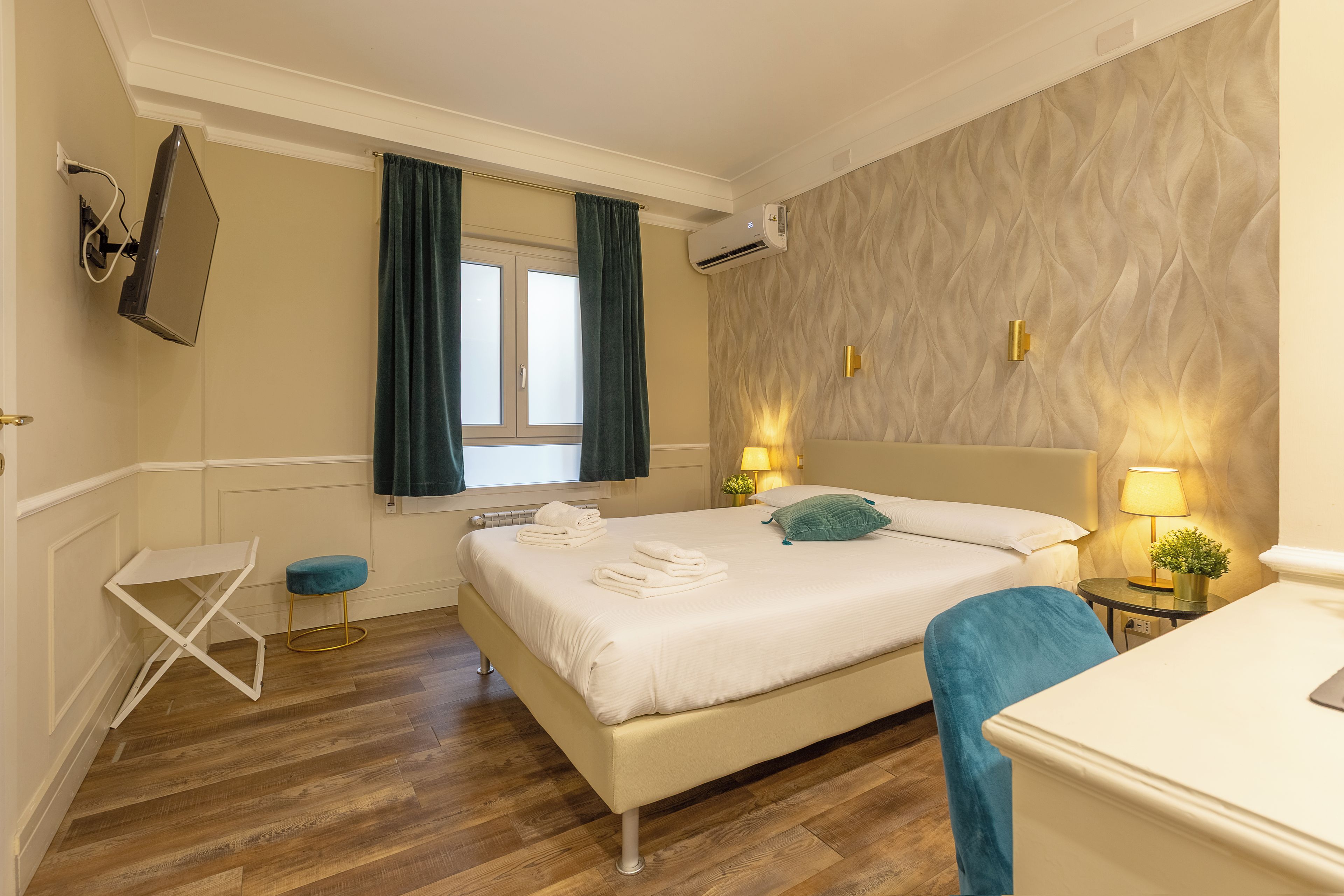 A hotel room with a bed, a small table, a chair, a window with curtains, an air conditioning unit, and a wall-mounted lamp.