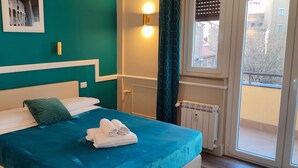 A neatly made bed with a teal bedspread, white pillows, and a green throw pillow. There are folded towels on the bed. A framed picture hangs on the teal wall above the bed. - Le Petit Bijou (Rome)