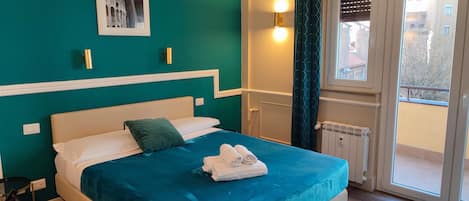 A neatly made bed with a teal bedspread, white pillows, and a green throw pillow. There are folded towels on the bed. A framed picture hangs on the teal wall above the bed.