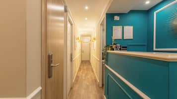 Corredor de hotel com portas de madeira, balcão de recepção e parede verde-água com quadros de certificados.