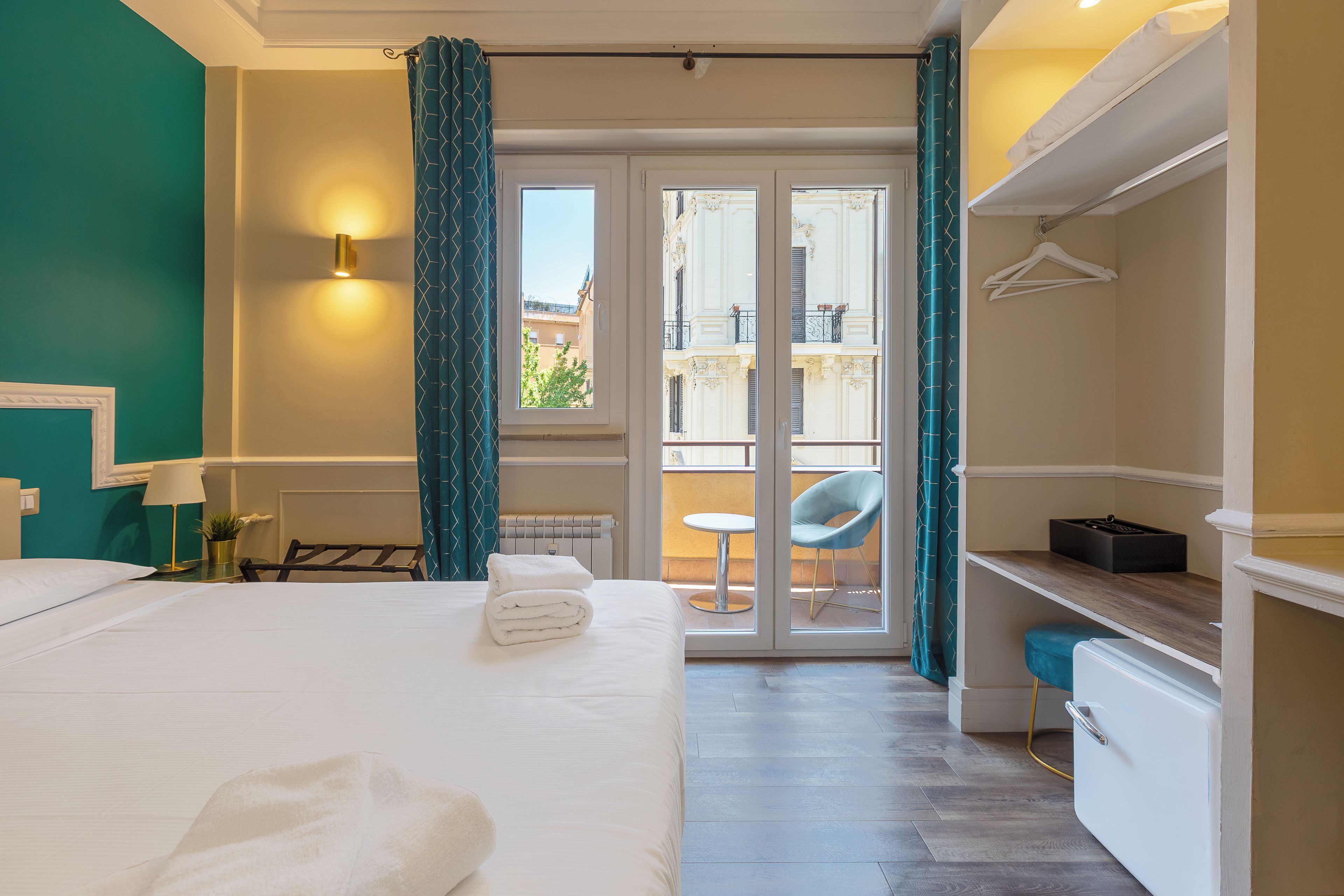 Quarto de hotel com uma cama, uma escrivaninha, uma cadeira, um radiador e vista através de uma porta de vidro.