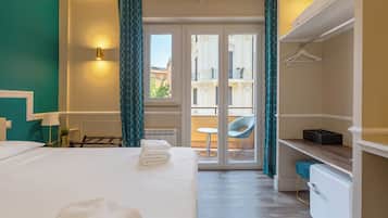 Quarto de hotel com uma cama, uma escrivaninha, uma cadeira, um radiador e vista através de uma porta de vidro.