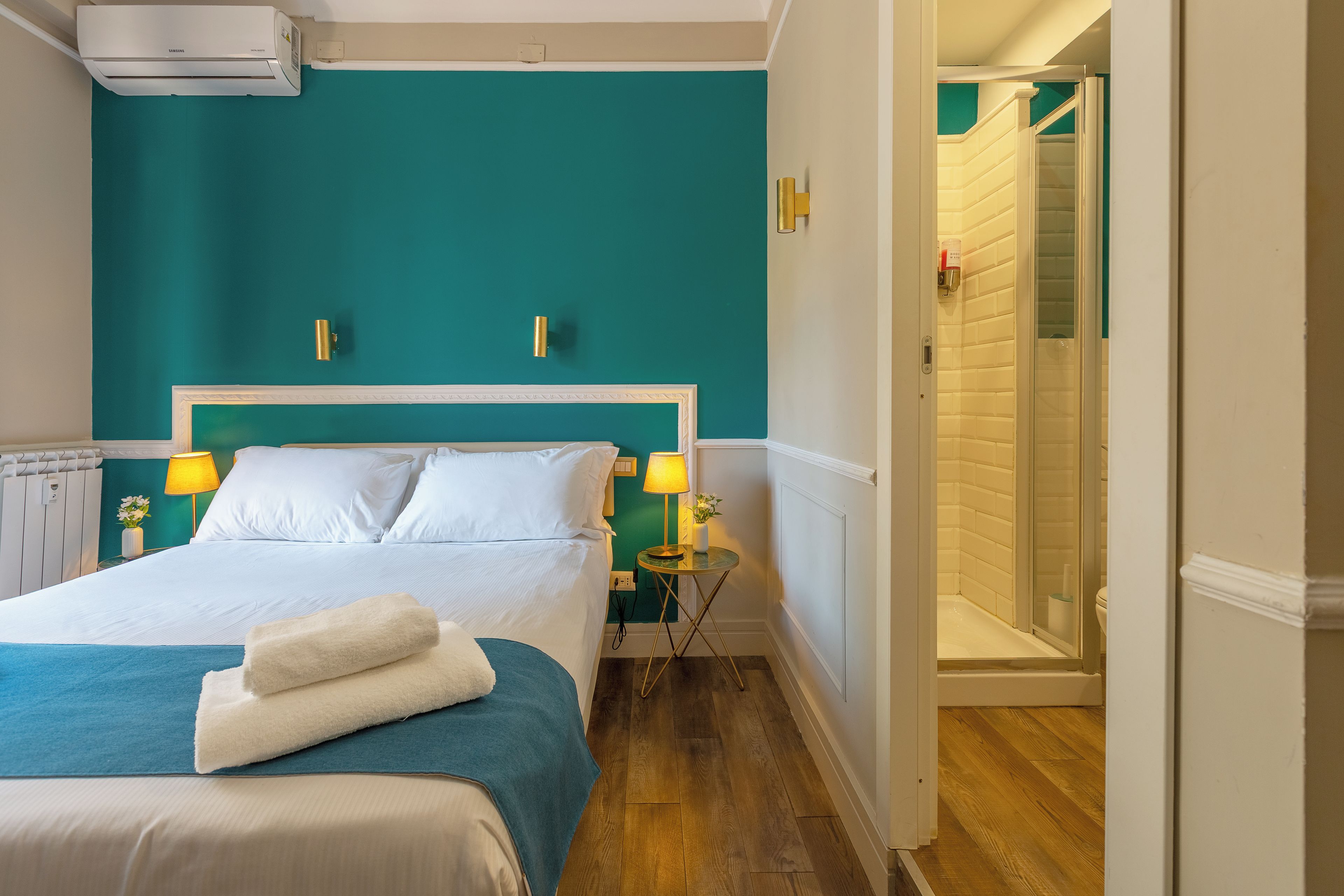 Habitación de hotel con cama, lámparas de noche, baño con ducha y pared color aguamarina.