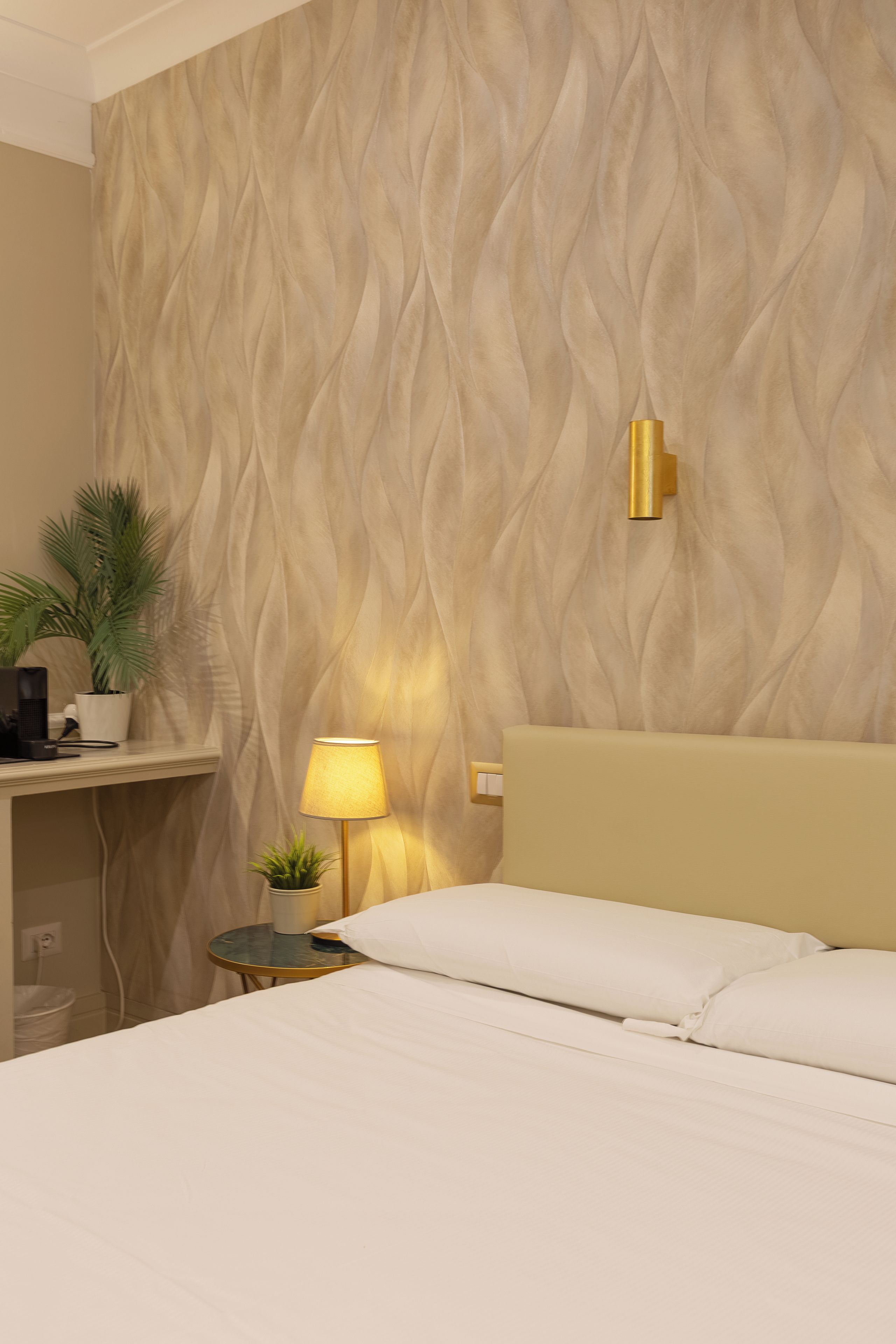 A hotel room with a bed, a bedside lamp, a small table with a plant, and a wall with a textured pattern.