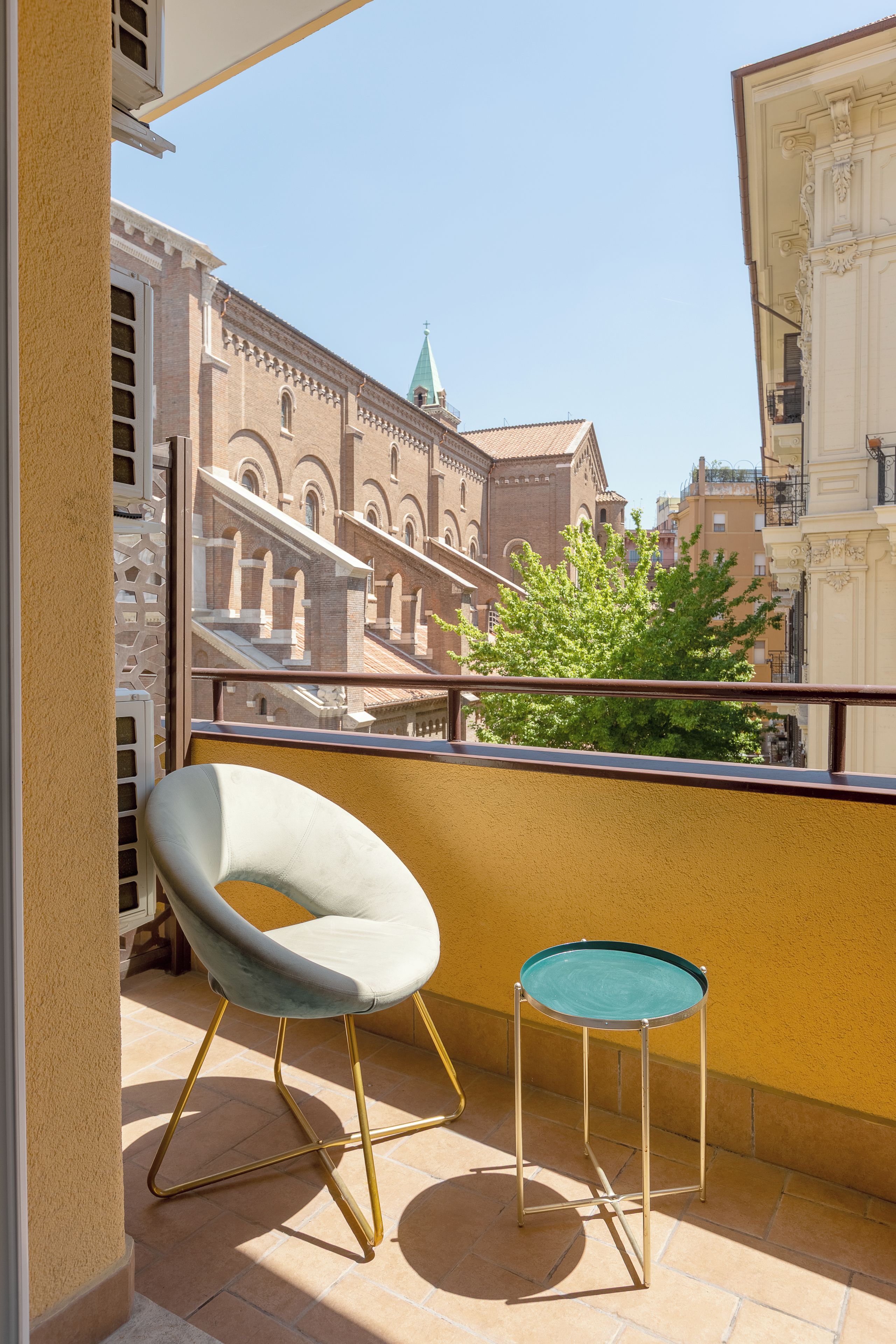 Varanda com cadeira e mesa pequena, vista para um edifício histórico com arcos e telhado verde.