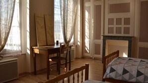 A room with a wooden desk, a chair, a bed with a patterned bedspread, a fireplace, and large windows with curtains.