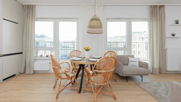 A modern living room with a dining area, featuring wicker chairs, a round table, and a hanging light fixture.