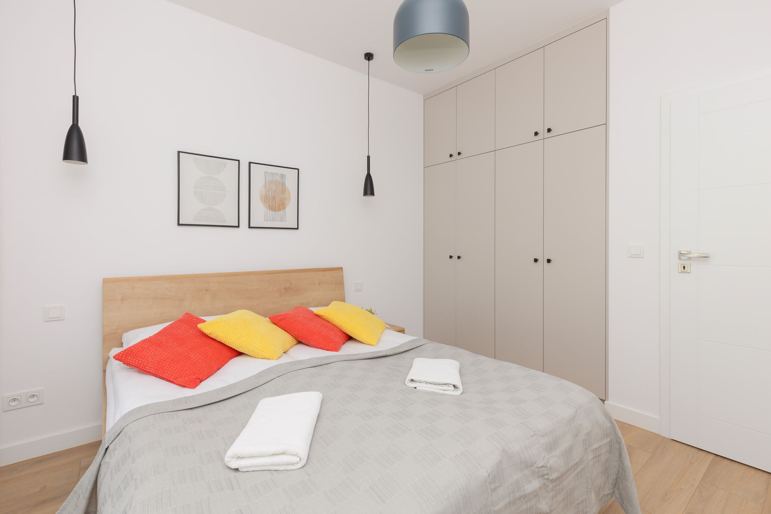 A neatly made bed with a grey bedspread, red and yellow pillows, and a white towel. There are two black pendant lights above the bed, and a large wardrobe with sliding doors is visible to the right.