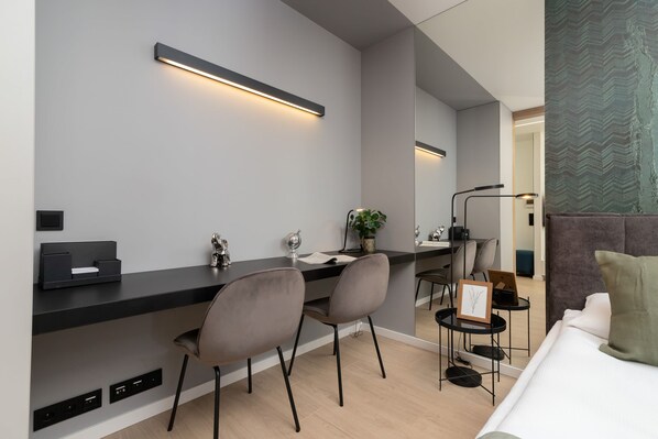 Habitación de hotel moderna con un escritorio gris oscuro, dos sillas grises, una mesita de noche con una planta, un cuadro enmarcado y una cama con almohada verde.