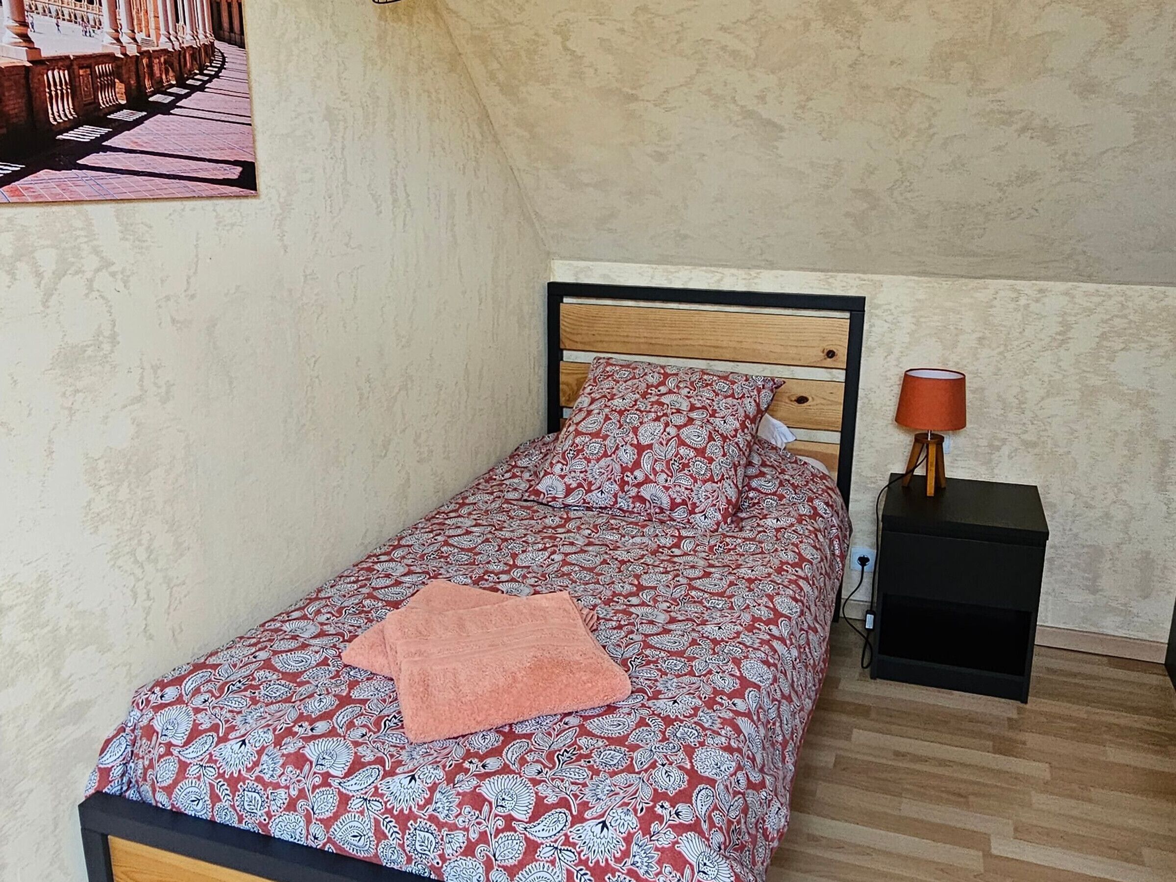 Un lit simple avec une couvre-lit à motifs et une tête de lit en bois, une table de chevet avec une lampe et un mur à texture.