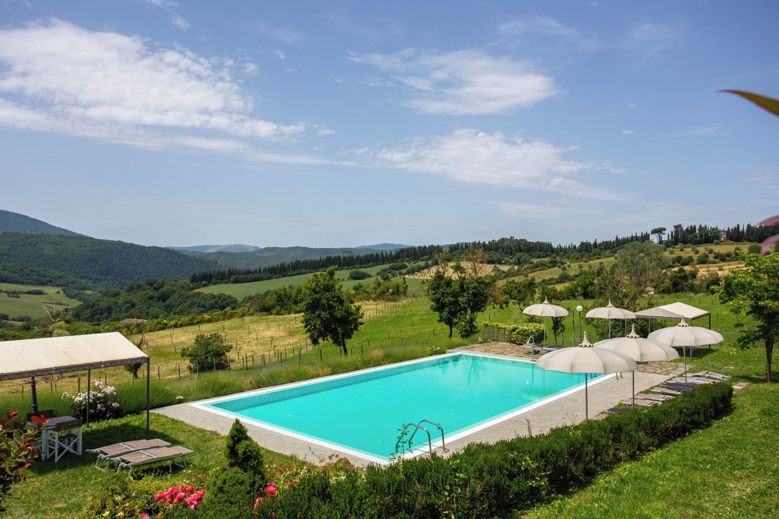 A rectangular swimming pool with a clear blue water surface, surrounded by a grassy area with several umbrellas and lounge chairs.