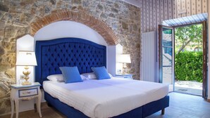 Een slaapkamer met een stenen muur, een groot bed met een blauw hoofdeinde, twee nachtkastjes met lampen, en uitzicht door een geopende deur.
