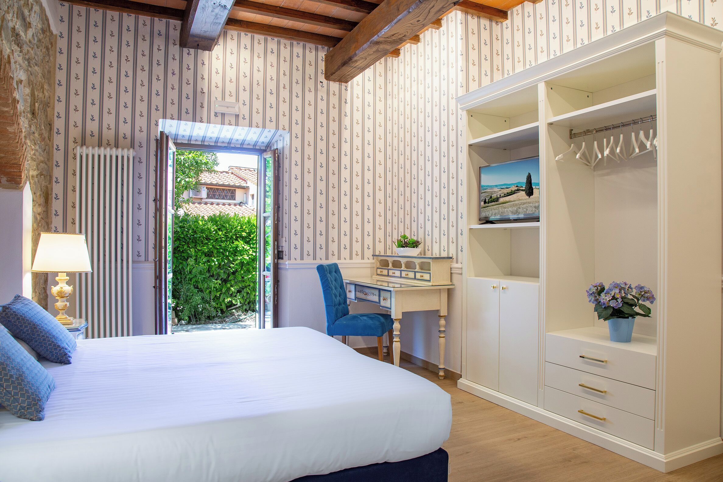 A bedroom with a bed, a desk, a chair, a wardrobe, and a view of greenery outside.
