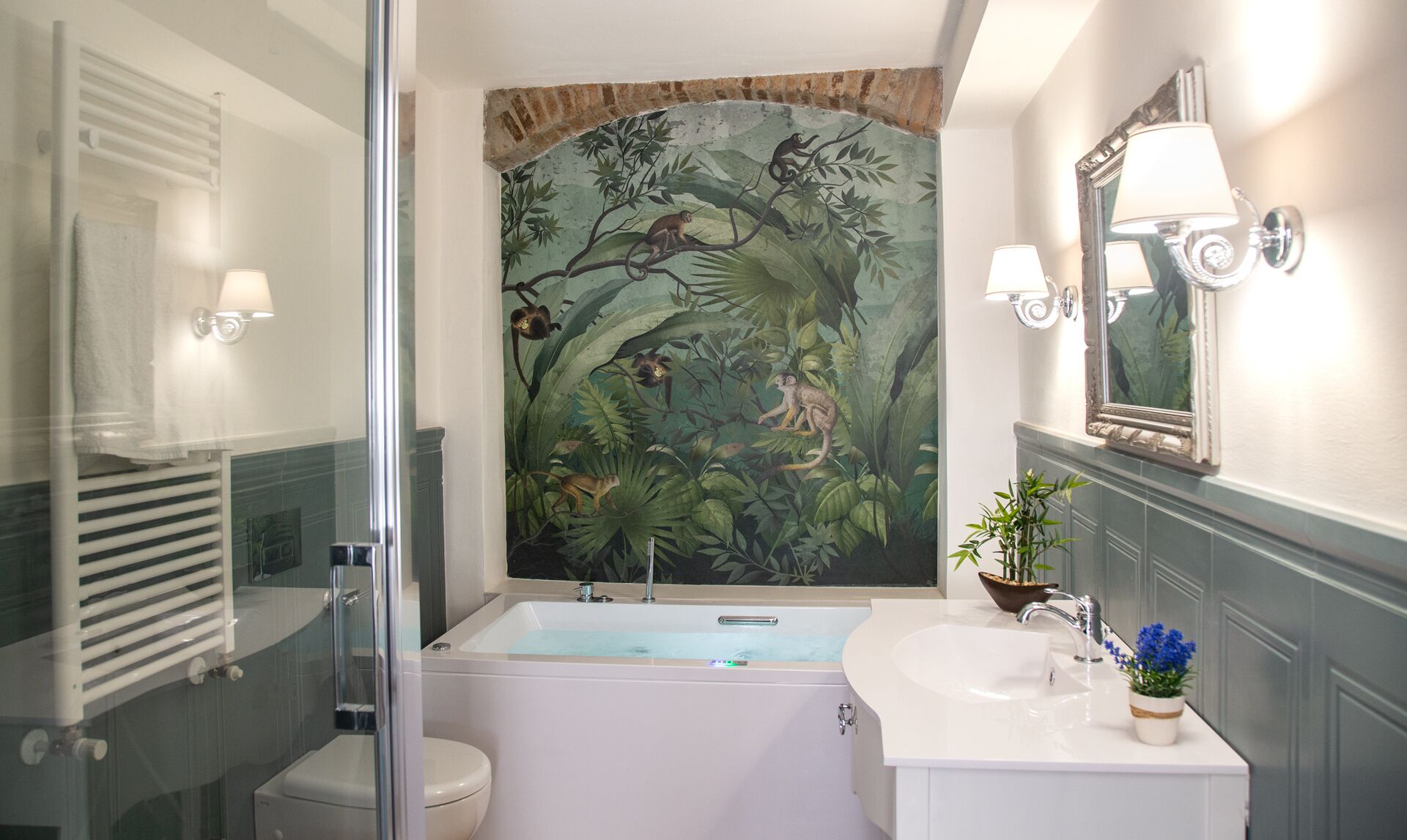 Un baño con bañera, inodoro, cabina de ducha de vidrio, lavamanos con espejo y un mural en la pared con monos y follaje.