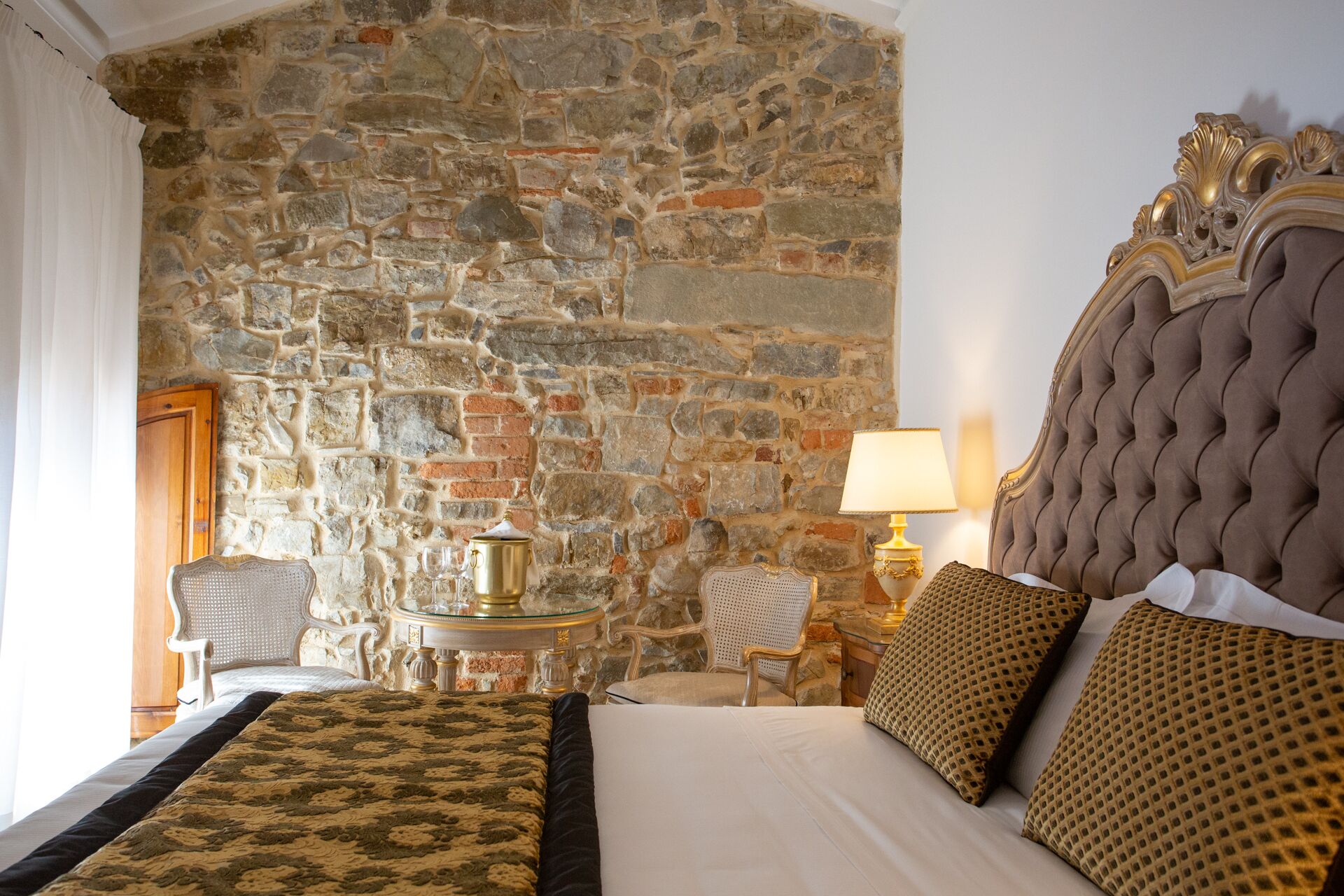 A bedroom with a stone wall, a bed with a tufted headboard, a nightstand with a lamp, and a chair.