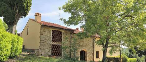 A stone building with a tiled roof, surrounded by greenery and a gravel path.