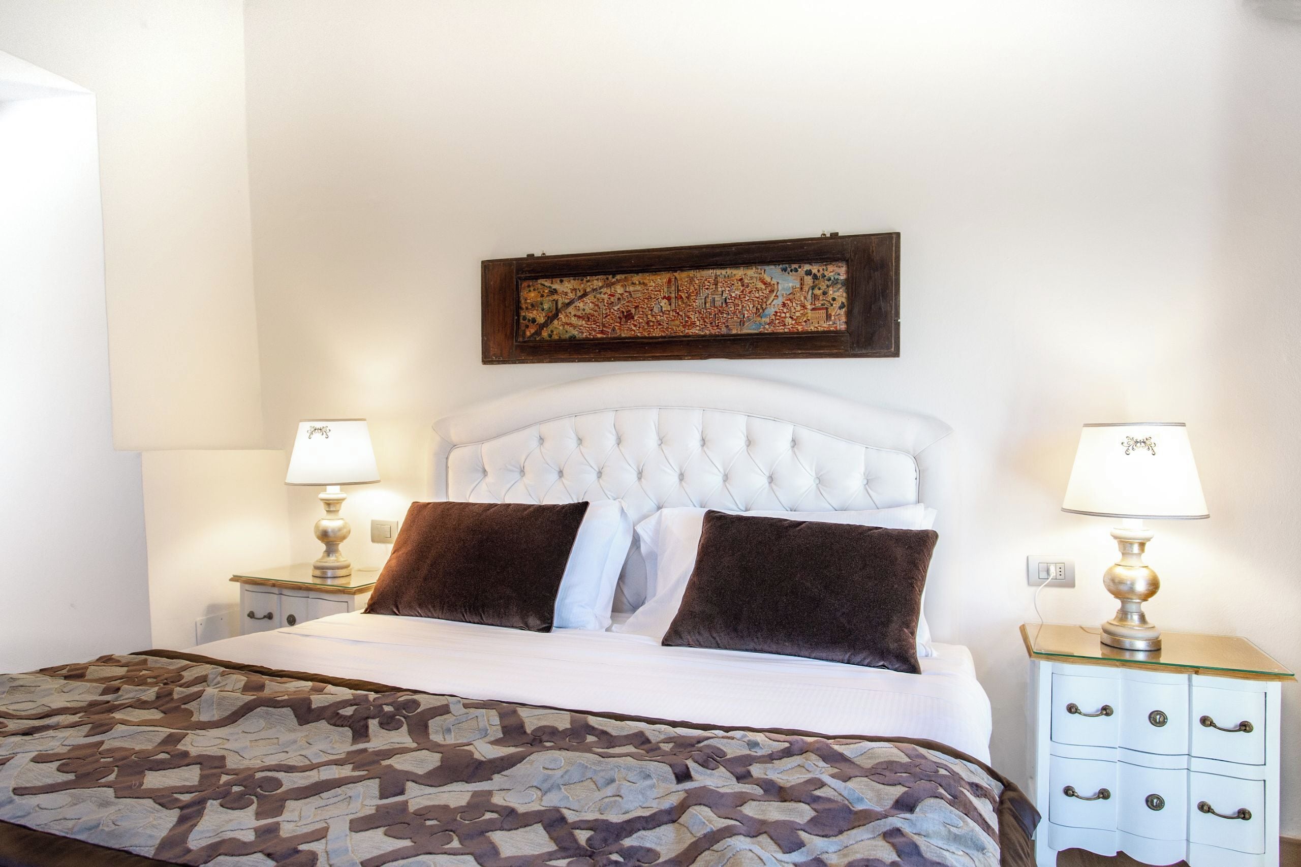 Una cama bien hecha con colcha estampada, cabecera acolchada, dos mesitas de noche con lámparas y una obra enmarcada sobre la cama.