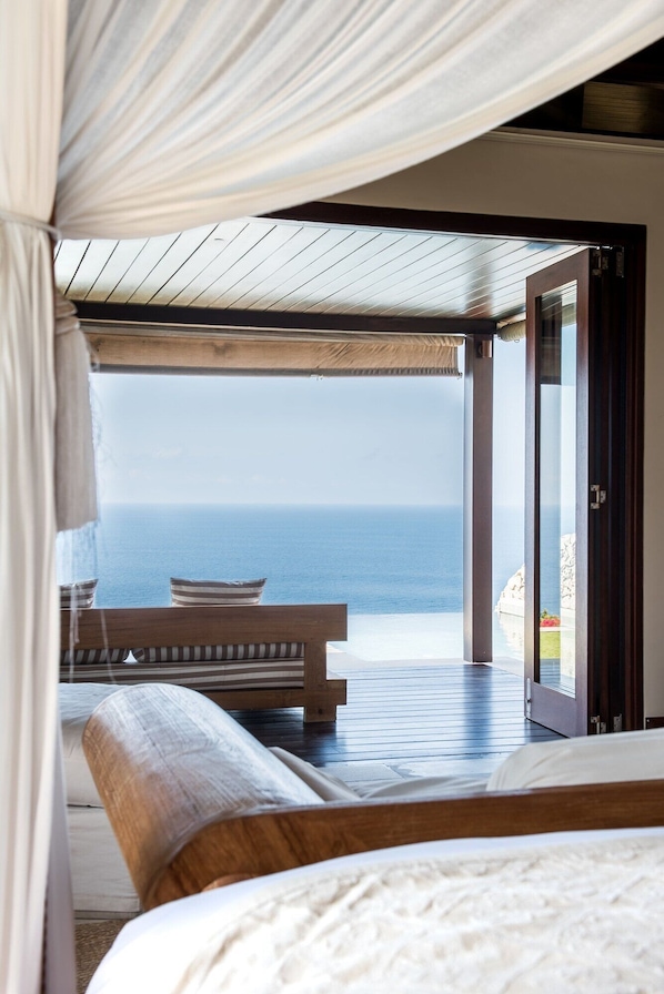 A bedroom with a large bed, a wooden bench, and a view of the sea through an open door. - The Ungasan Clifftop Resort (Pecatu)