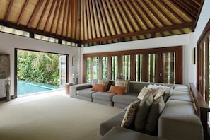 A spacious living room with a large sofa, a pool, and a view of lush greenery. - The Ungasan Clifftop Resort (Pecatu)