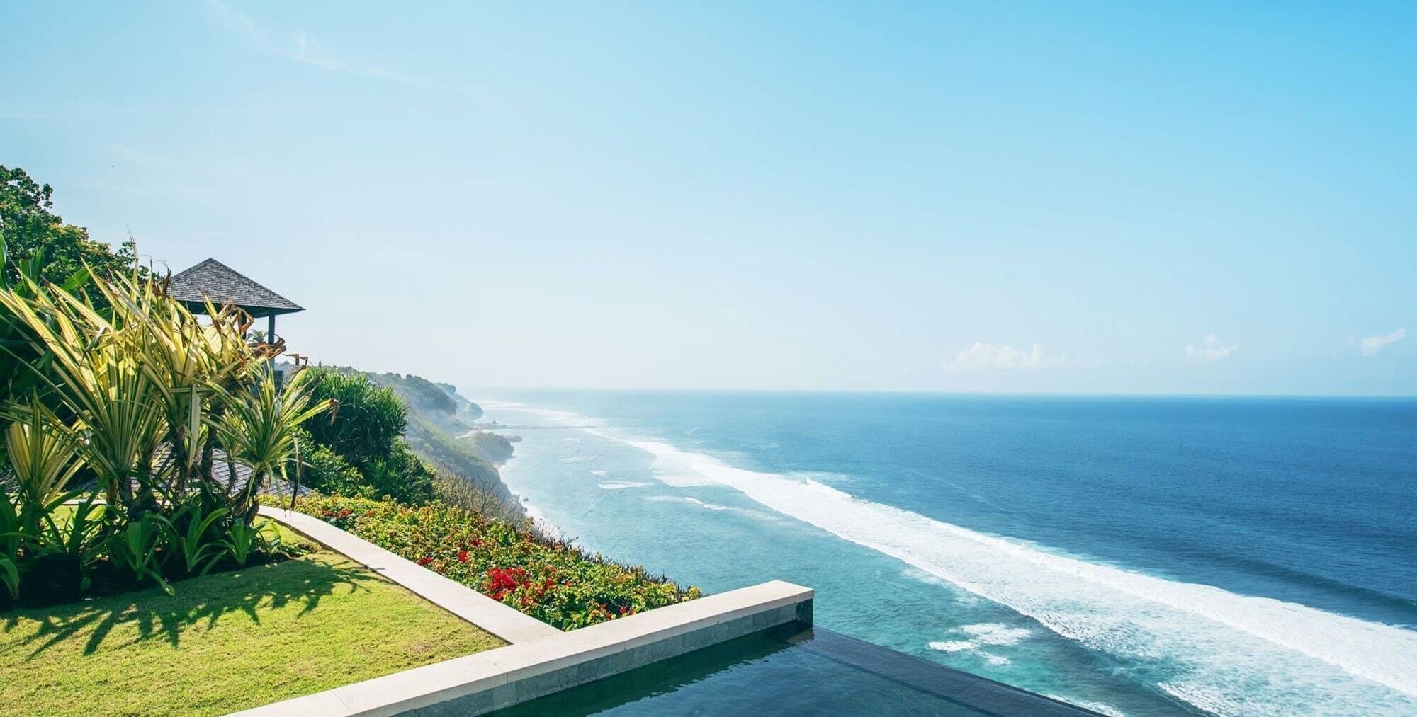 A coastal view with a pool, a thatched-roof structure, and lush greenery.