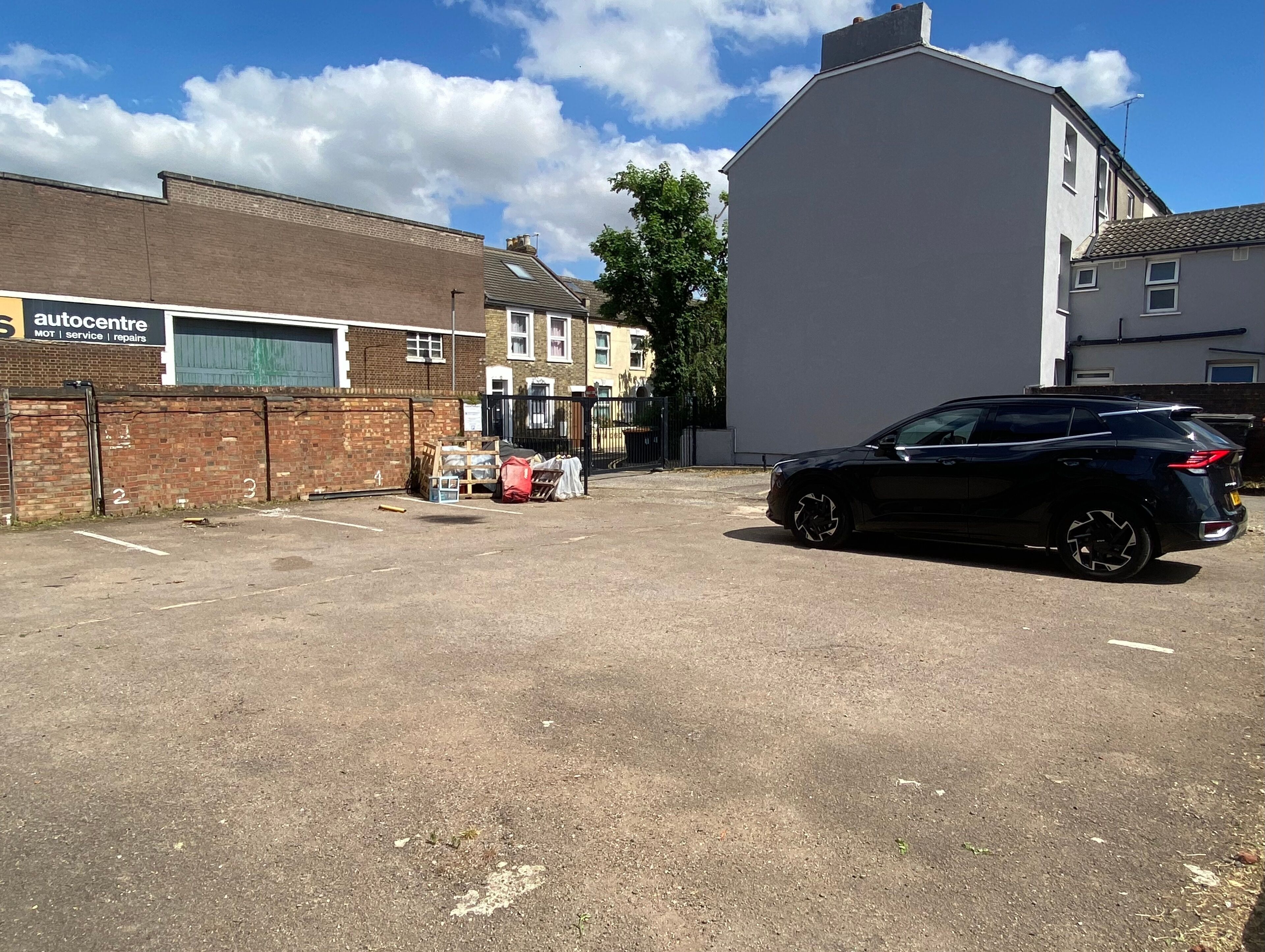 Ein Parkplatz mit einem schwarzen Auto auf der rechten Seite, einem Gebäude mit der Aufschrift 'autocentre' auf der linken Seite und blauem Himmel.