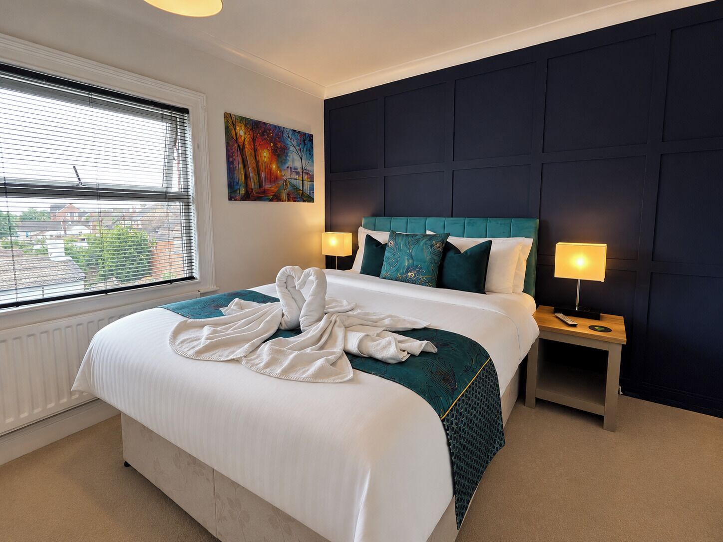 A hotel room with a bed, two bedside lamps, a painting on the wall, and a window with blinds.